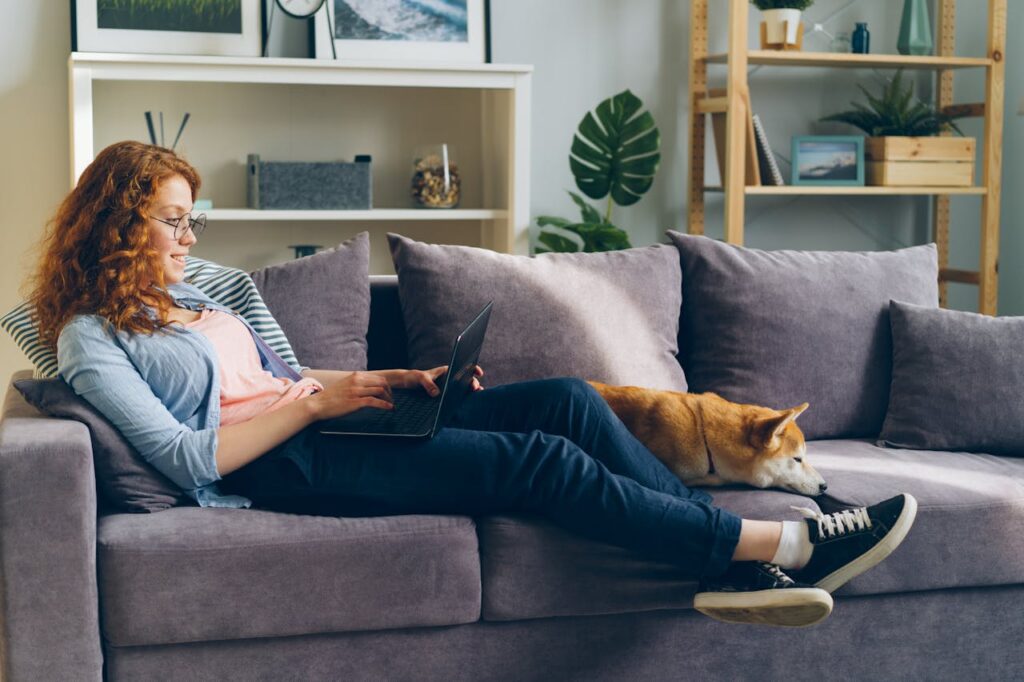 A woman sitting on a couch holding a laptop while her dog lays next to her