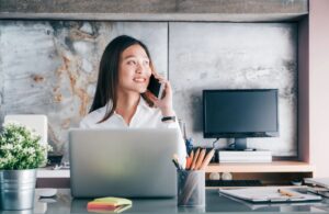 woman sitting at a desk on her phone with a laptop in front of her